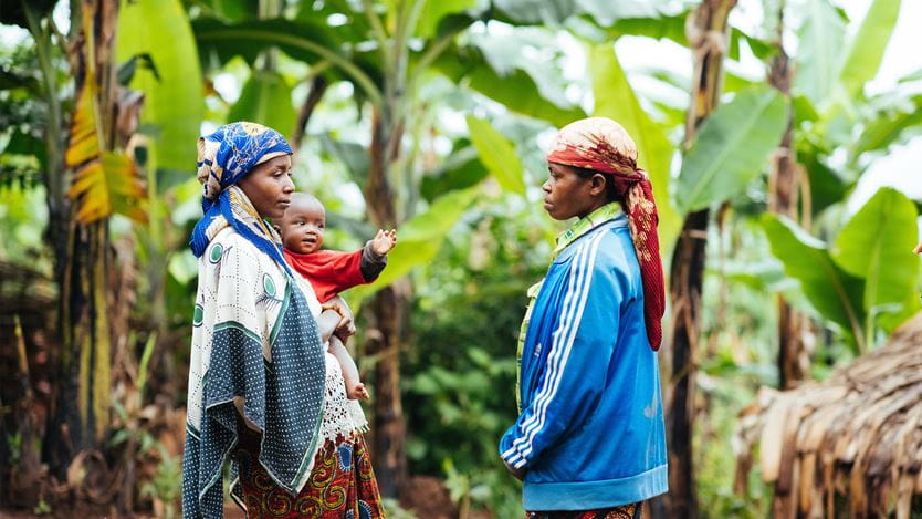 Duas mulheres, uma delas segurando uma criança, de pé em uma plantação de bananeiras, participando de um programa de nutrição alimentar