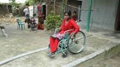 Uma senhora, vestindo um sari vermelho, andando de cadeira de rodas perto de uma casa