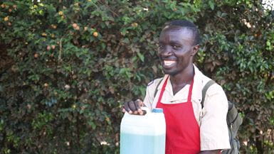 Um homem chamado Festus, vestindo um avental vermelho, sorrindo e segurando um recipiente de plástico com o sabão líquido que ele vende em seu negócio