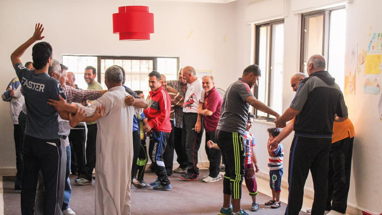 Hombres jóvenes y mayores de una comunidad se reúnen en círculo, algunos abrazándose, bailando y moviéndose como parte de una terapia de trauma y recuperación