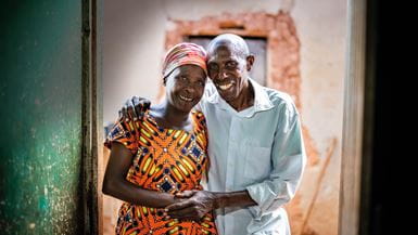 Un couple africain âgé se tient la main en souriant à l’entrée de sa maison.