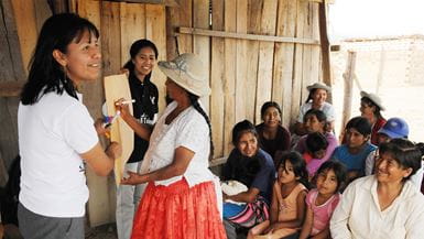 Na zona rural da Bolívia, duas mulheres sorrindo seguram um cartaz para outra mulher escrever, enquanto um grupo de mulheres e crianças assistem