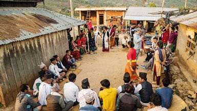 A large number of Nepalese women and men sit on the ground in a circle to watch a group of actors perform
