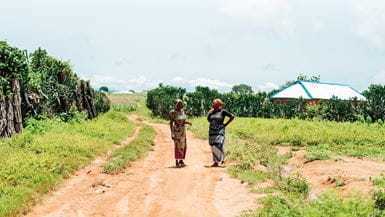 Dos mujeres nigerianas conversan de pie en una calle de tierra rural con un edificio en la distancia.