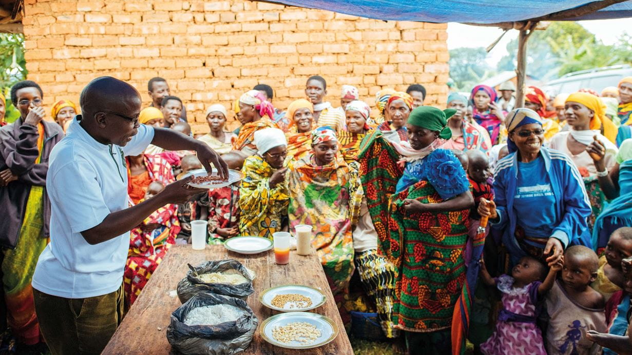 Um homem do Burundi sendo observado por um grupo de mulheres e crianças enquanto segura um prato de grãos sobre sacos e pratos com diferentes tipos de farinha, feijões e grãos em cima de uma mesa de madeira
