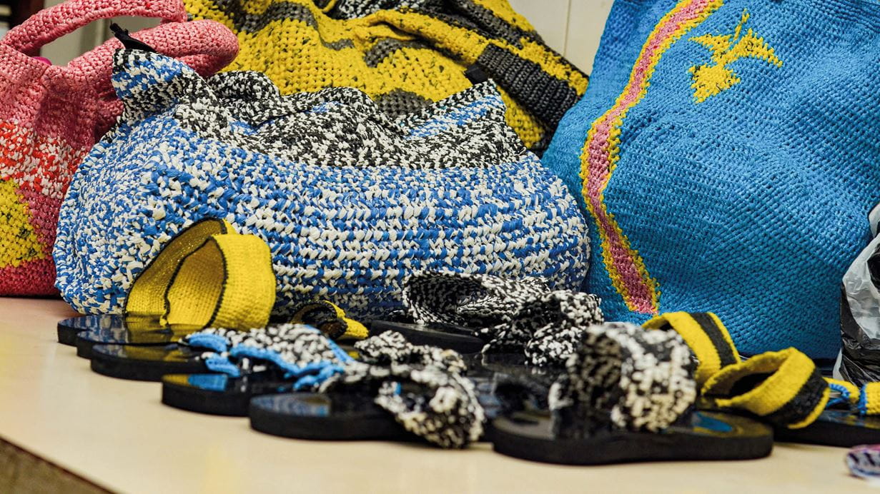 Sacolas estampadas azuis, rosas e amarelas tramadas com resíduos plásticos ao lado de sandálias coloridas