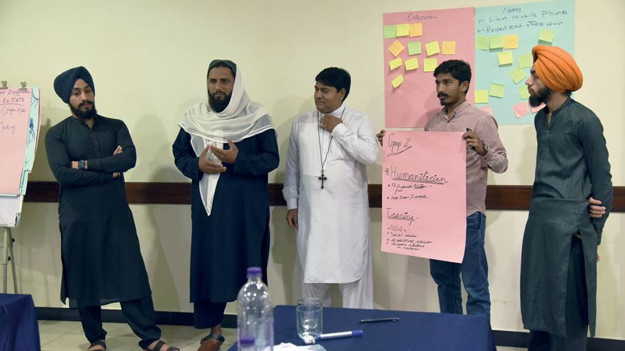 Cinq hommes pakistanais de religions différentes se tiennent côte à côte dans une salle, l'un d'entre eux montrant une grande feuille de papier rose avec des inscriptions