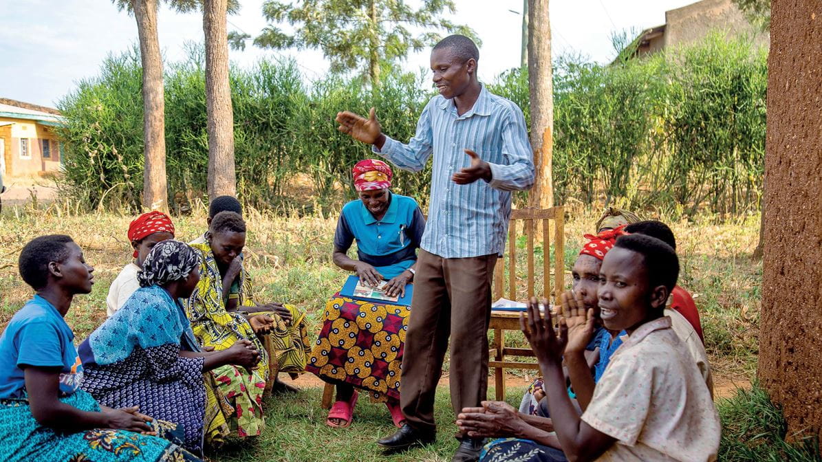 Au Burundi, un homme souriant se tient debout au milieu d'un groupe de femmes assises, vêtues d'habits aux couleurs vives