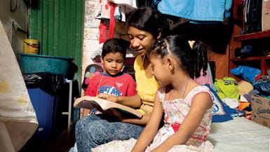 Uma mulher, um menino e uma menina sentados em uma cama em sua casa na Colômbia, olhando juntos para uma Bíblia