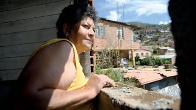 Uma mulher colombiana de camiseta amarela, olhando pela janela para casas feitas de tijolos e com telhados de zinco