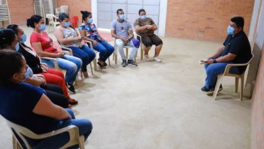 A group of men and women sit in a semi-circle on plastic chairs as a man wearing a black polo leads them in a devotional session.