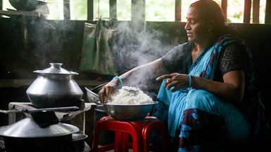 A woman wearing a bright blue sari dress sits on the ground and cooks steaming hot white rice in a cast iron bowl for her family.