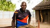 Un pasteur du Soudan du Sud œuvrant à la consolidation de la paix est assis sur un banc en métal devant une hutte en paille, un long crucifix en bois à la main.
