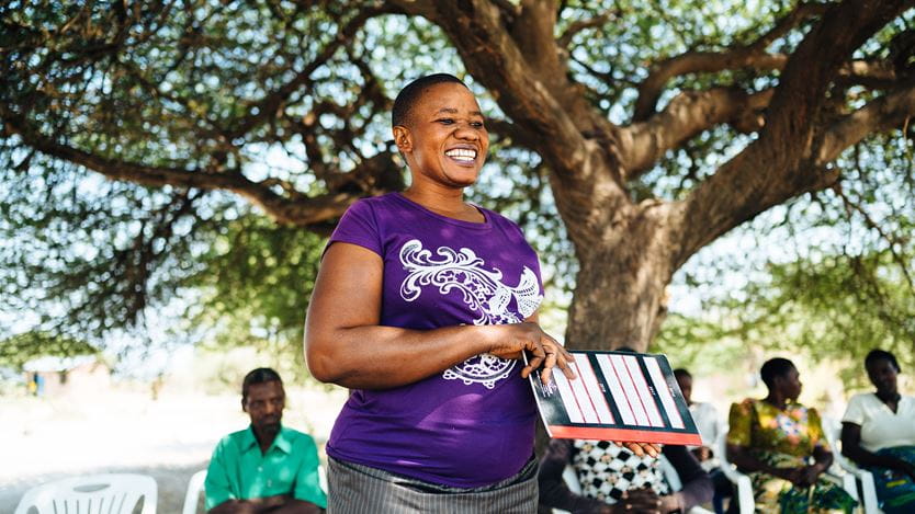 A smiling African woman in a purple tope speaking to community members gathered under a tree
