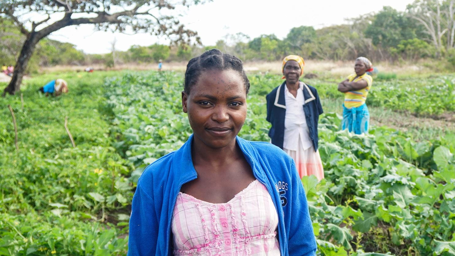 Uma mulher em pé sorrindo entre plantações, em um campo em Moçambique, com duas mulheres no fundo, à distância