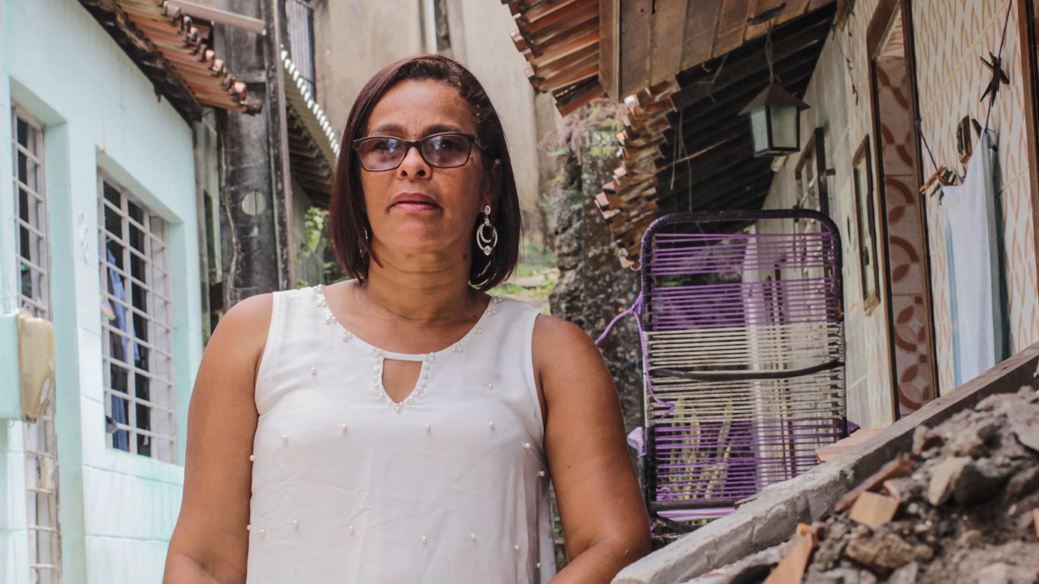 Une militante se tient debout devant des maisons d’une rue étroite du Brésil, une chaise longue en métal à côté d’elle.
