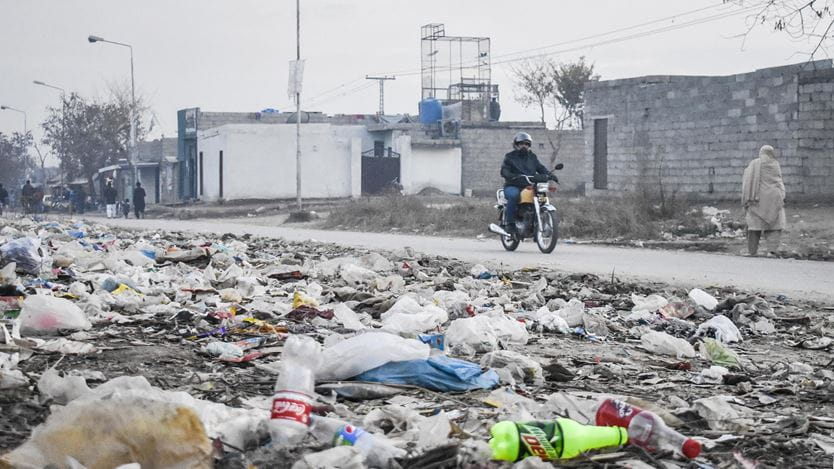 Un homme à moto suit un chemin bordé d’un haut mur de briques grises et jonché de bouteilles en plastique de Coca-Cola.