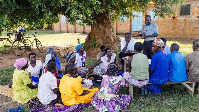 Homens e mulheres ugandenses sentados em um banco sob uma árvore, em uma reunião comunitária