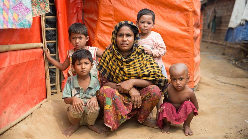 Una mujer posa en cuclillas sobre un suelo polvoriento, rodeada de sus cuatro hijos pequeños fuera de su carpa en un campamento de refugiados rohingya