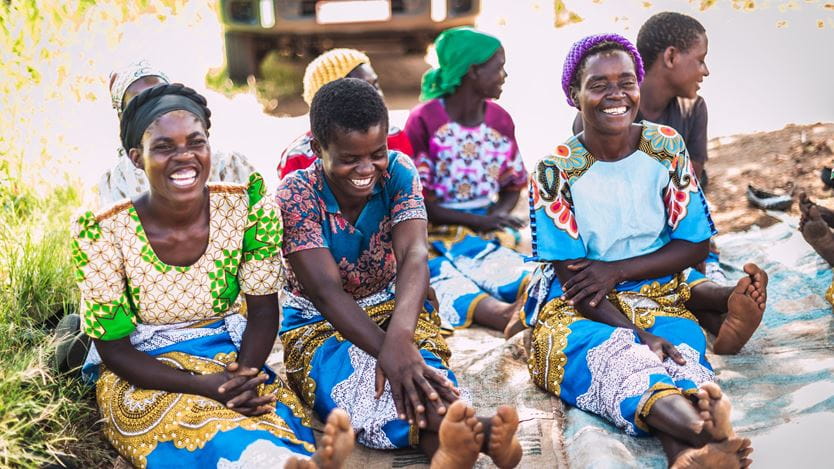 A group of smiling people are sitting on a cloth on the ground outdoors, with a vehicle partially visible in the background. The people in the foreground are barefoot and wearing brightly coloured, patterned clothing.