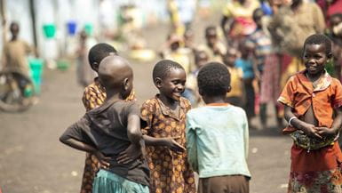 A group of young children are gathered together outdoors, smiling and interacting with one another. The children in the foreground are in clear focus, while a large, diverse crowd of people and a line of colorful containers are visible in the blurred background.