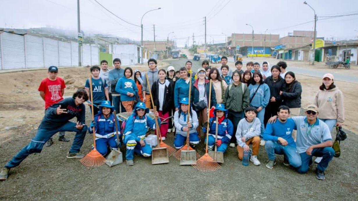 A large group of people, including adults and young people, pose on a dusty road. Several wear blue uniforms and hats, holding rakes and dustpans. Buildings and a street are visible in the background under a cloudy sky.
