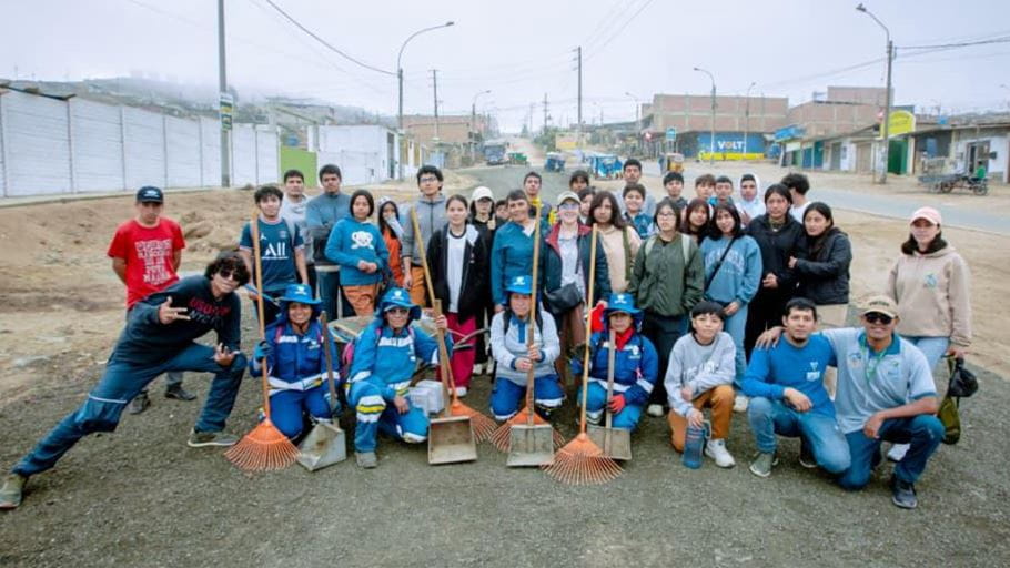 A large group of people, including adults and young people, pose on a dusty road. Several wear blue uniforms and hats, holding rakes and dustpans. Buildings and a street are visible in the background under a cloudy sky.