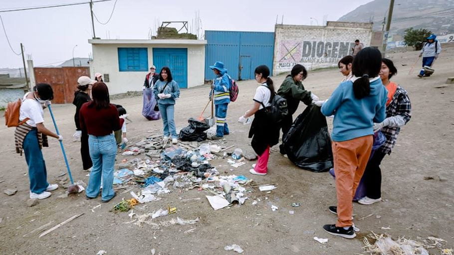 A group of around a dozen young people, wearing gloves, are collecting litter from a dusty, unpaved area. Some are sweeping rubbish into piles, others hold black bin bags. Buildings and hills are visible in the background.