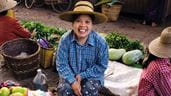 Uma mulher sentada no chão, usando um chapéu de palha e sorrindo enquanto vende enguias e outros peixes em Hsipaw, em Myanmar