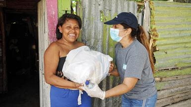 Duas mulheres de pé, uma ao lado da outra, na frente de um barraco de ferro corrugado na Colômbia, uma delas entregando um pacote de alimentos como parte da resposta à Covid-19