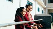 Deux femmes se tiennent la main sous un robinet commun dans leur école au Népal, en se souriant l’une à l’autre.