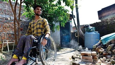 Un homme sourit, assis dans un fauteuil roulant devant un bâtiment à côté d’une poubelle bleue et d’une pile de briques.