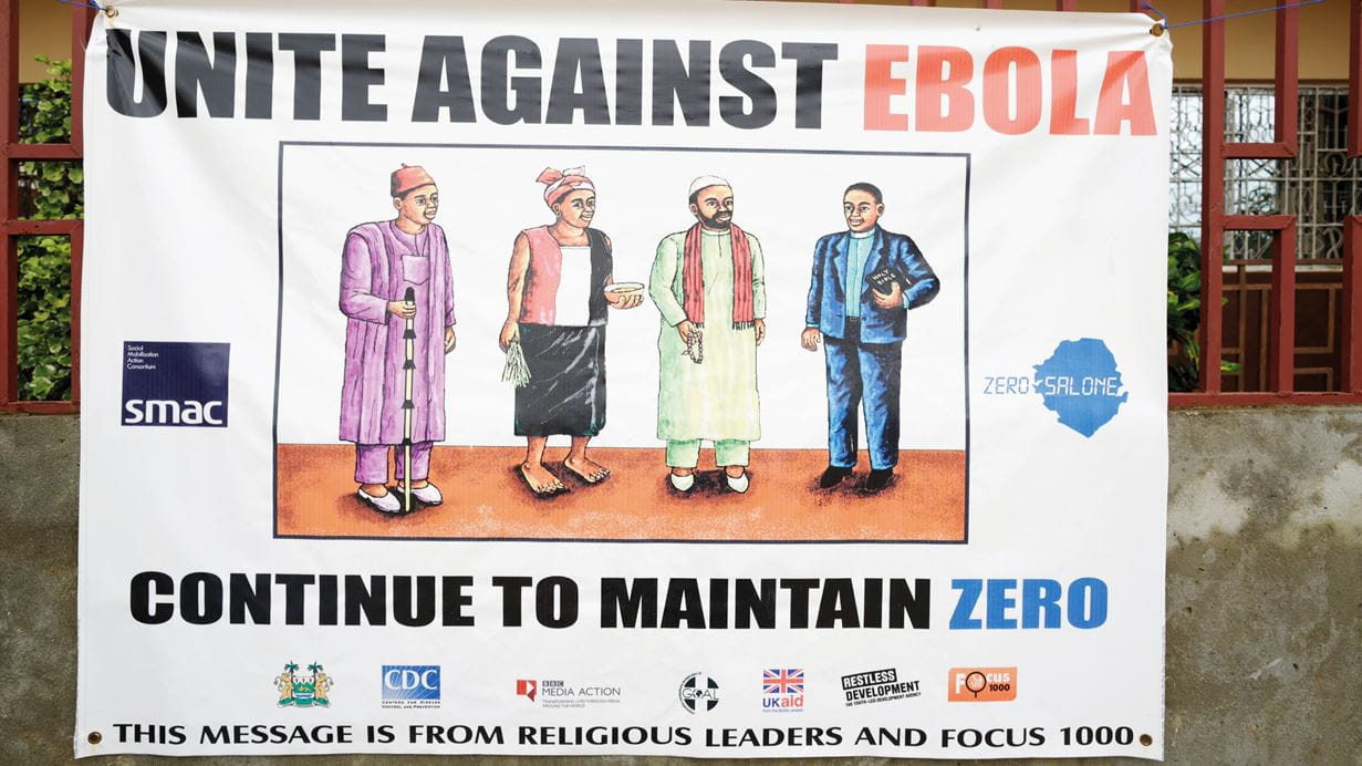 Un enorme cartel fuera de una construcción en Sierra Leona ilustra un dibujo de cuatro líderes de fe y la leyenda «Unite against Ebola. Continue to maintain zero» (Unidos contra el ébola. Continuemos en cero)