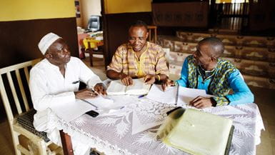 Tres hombres líderes de fe con la vestimenta tradicional de Sierra Leona, sentados junto a una mesa de madera, conversan sobre el brote del ébola