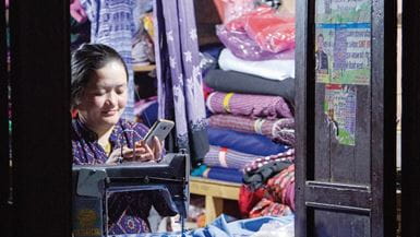Uma costureira no Nepal sentada em frente à sua máquina de costura, procurando notícias em seu telefone celular