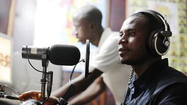 Un DJ de radio en Sierra Leona con audífonos habla frente al micrófono en una emisora de radio del organismo de desarrollo NEHADO