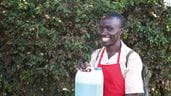 Um homem chamado Festus, vestindo um avental vermelho, sorrindo e segurando um recipiente de plástico com o sabão líquido que ele vende em seu negócio