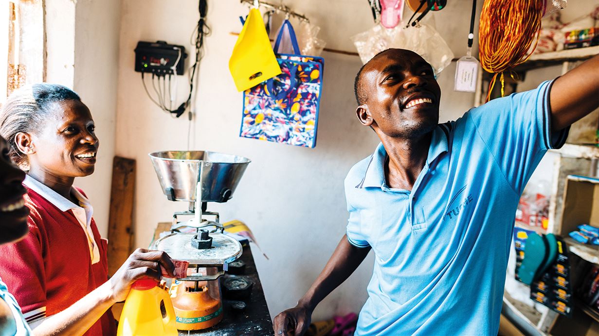 Lucas alcanza una repisa alta en su tienda de alimentos en Tanzania, mientras conversa y sonríe con una clienta