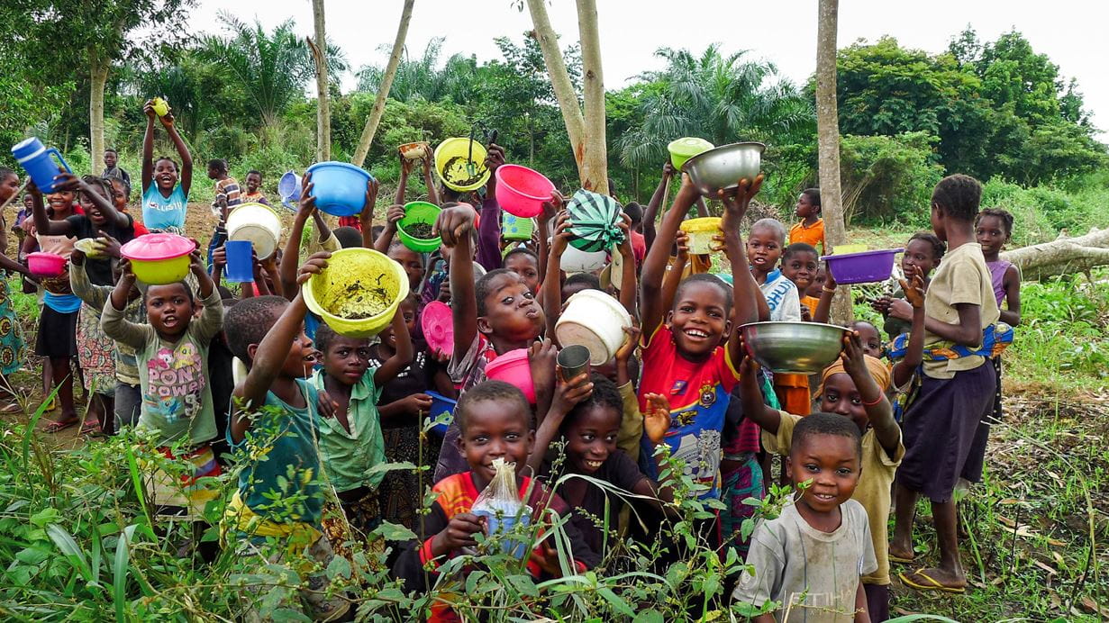 Um grupo grande de crianças sorridentes de pé no meio de uma floresta exuberante, segurando recipientes de plástico coloridos, usados para colher lagartas
