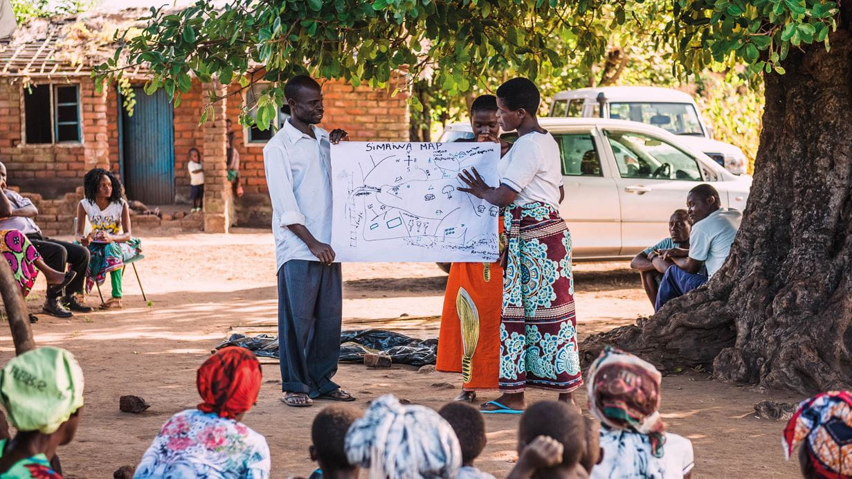 Une animatrice réalise un exercice de cartographie au Malawi devant un groupe de personnes à l’ombre d’un arbre.