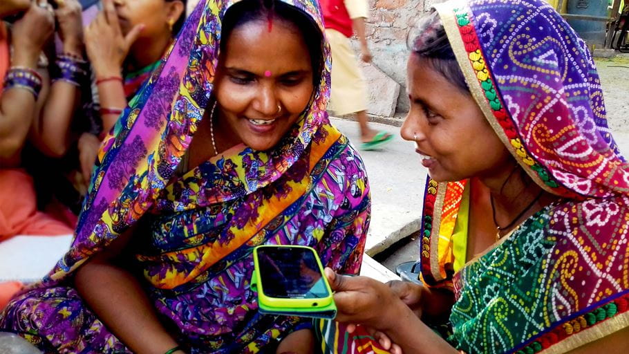 Two seated Indian women wearing colourful saris practise recording their stories on a a mobile phone
