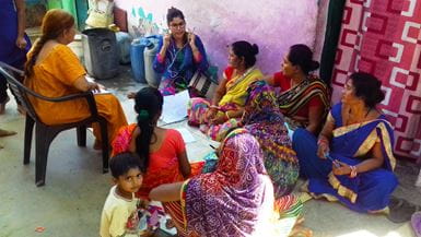 Um grupo de mulheres indianas sentadas assistem a outra mulher indiana demonstrando como usar um telefone celular para gravar histórias