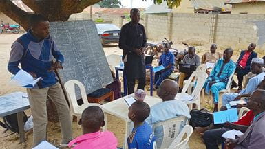 A group of seated Nigerian farmers listen to two Nigerian facilitators who are standing either side of a blackboard