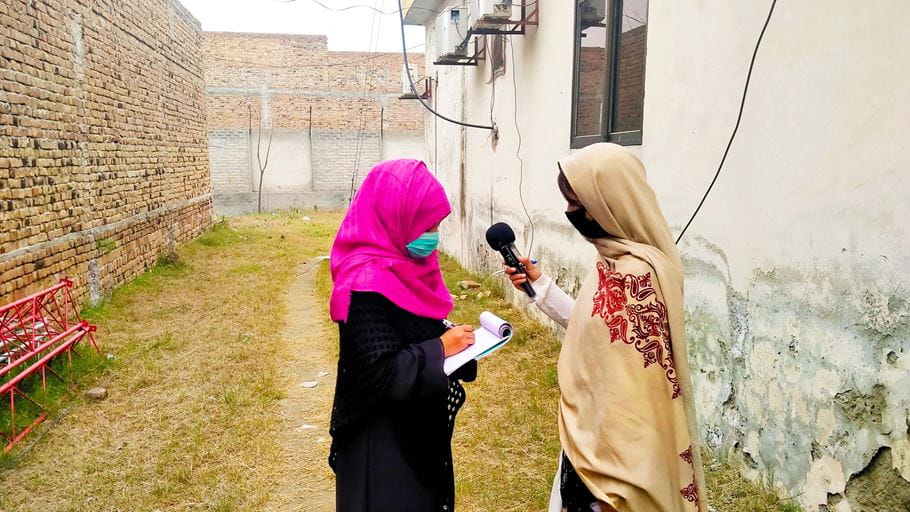 Two women wearing headscarves stand behind a house in Pakistan, and one speaks into a microphone