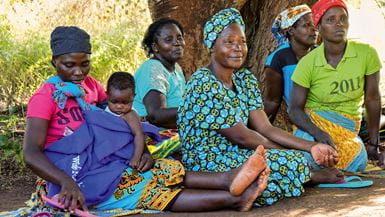 A group of Mozambican women, with one young child, sitting together on the ground under the shade of a tree