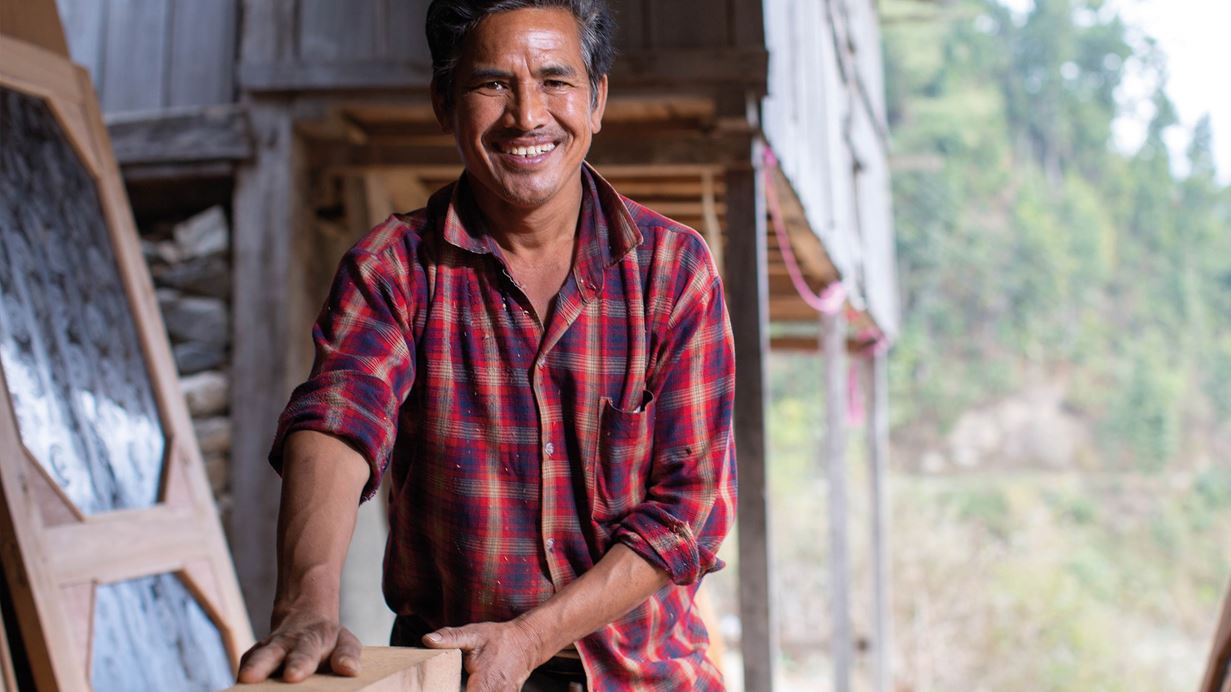 Um homem nepalês sorridente trabalhando com madeira em sua oficina em casa