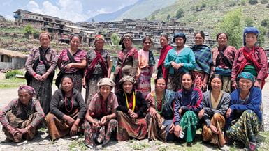 Um grupo de dezoito mulheres nepalesas posando para a câmera em um ambiente rural, com montanhas e um povoado no fundo