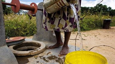 Primer plano de una mujer zimbabuense, descalza, de pie junto a un pozo, mientras vierte agua en un recipiente amarillo, grande, de plástico.