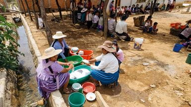 Tres mujeres con sombrero y una niña, en Bolivia, lavan los platos en palanganas de colores sentadas en cuclillas sobre el suelo polvoriento, cerca de otros miembros de la comunidad sentados a la sombra de unos árboles
