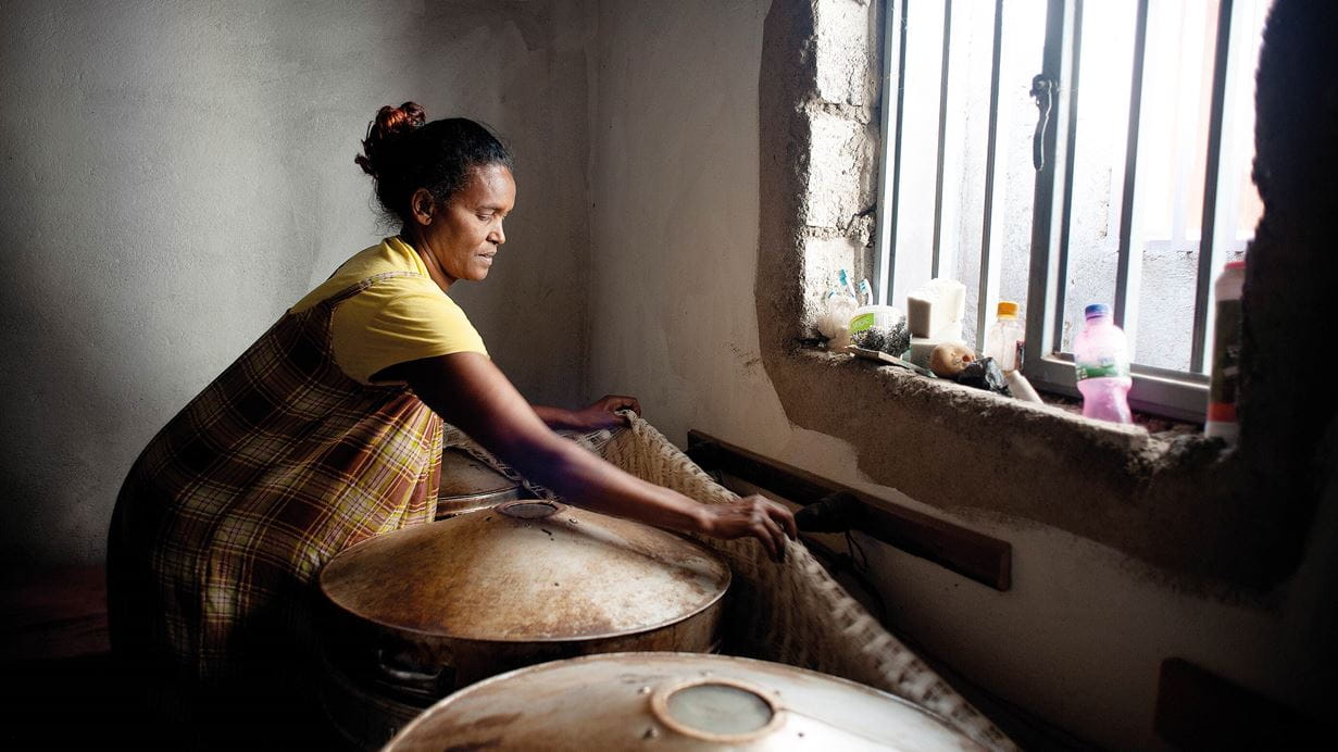 A female baker from Ethiopia wearing a yellow dress leans over towards a sunlit window and covers three large metal bread ovens with a cloth